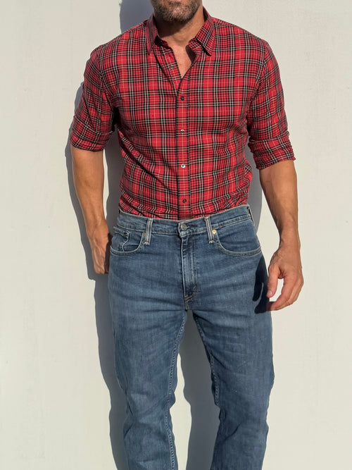 A man wearing a red and black plaid button-down shirt with rolled-up sleeves and blue jeans, standing against a plain light wall.