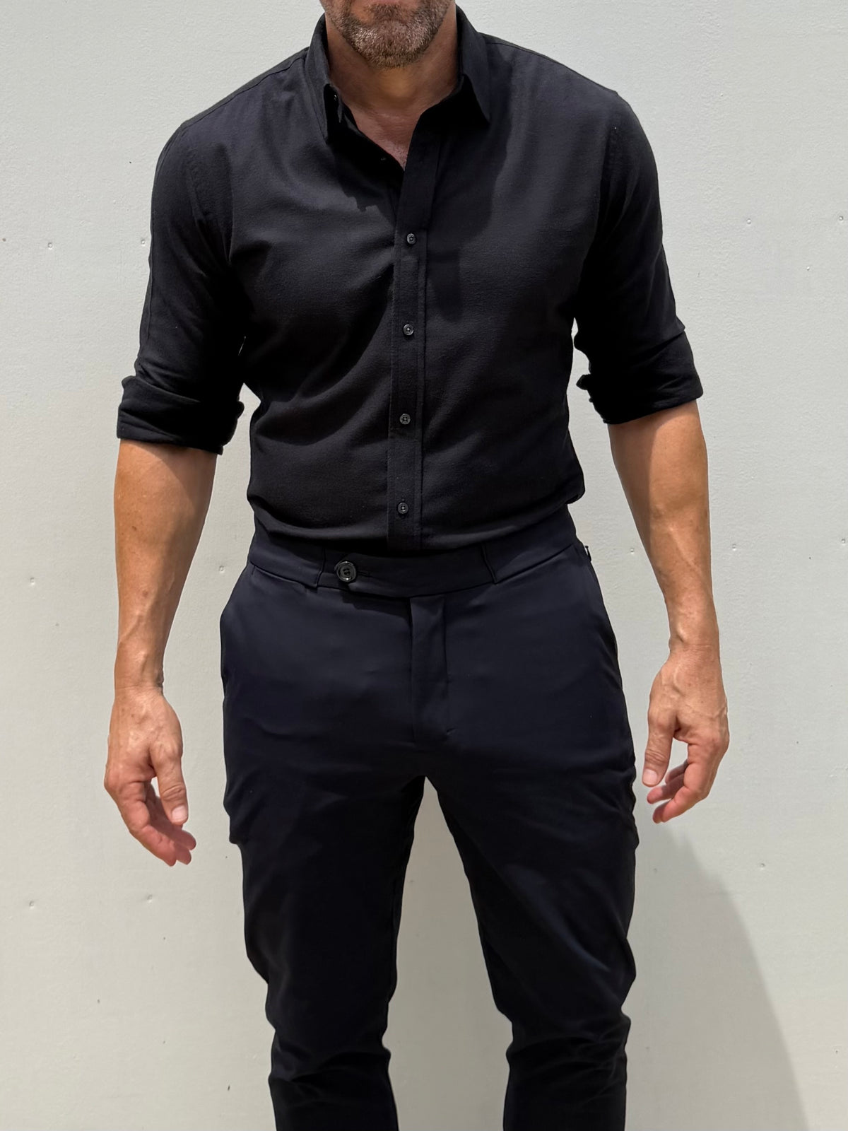 A man wearing a black long-sleeve button-down shirt with rolled-up sleeves and dark trousers, standing against a plain light gray wall.