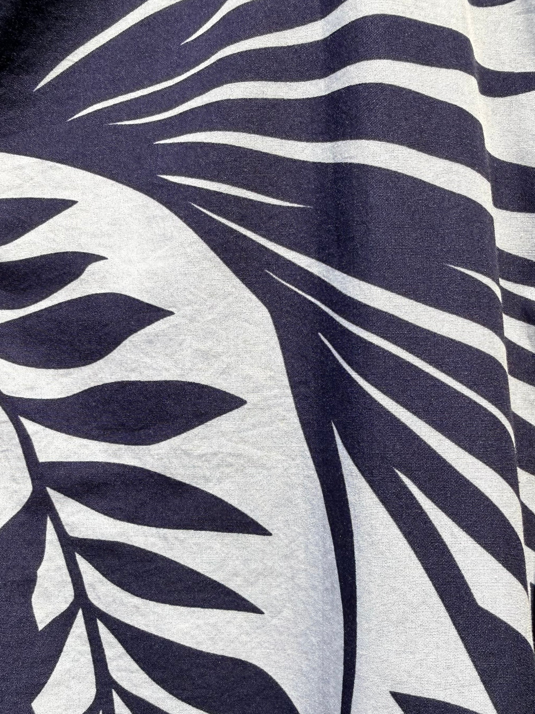 Close-up of a tropical leaf print fabric with dark navy blue palm fronds on a white background, showing part of a buttoned garment.