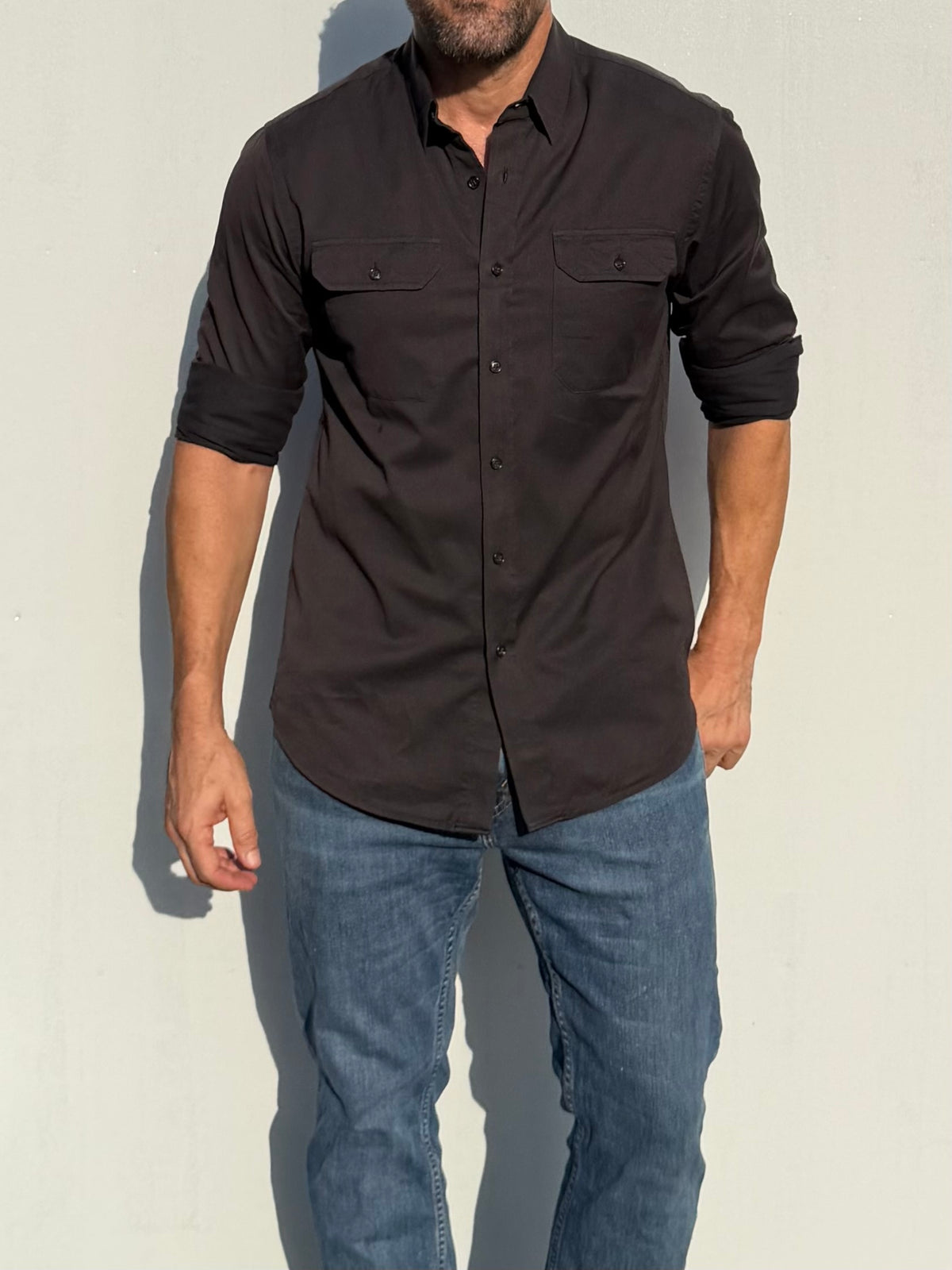 A man wearing a dark brown long-sleeve button-down shirt with chest flap pockets and blue jeans, standing against a light gray wall.