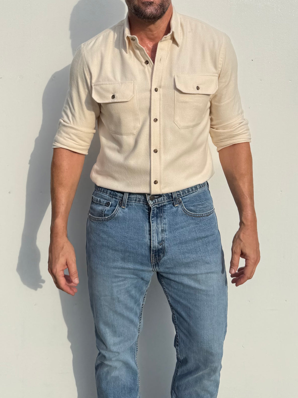 A man wearing a light beige long-sleeve button-up shirt with two chest pockets and light blue jeans, standing against a plain light wall.