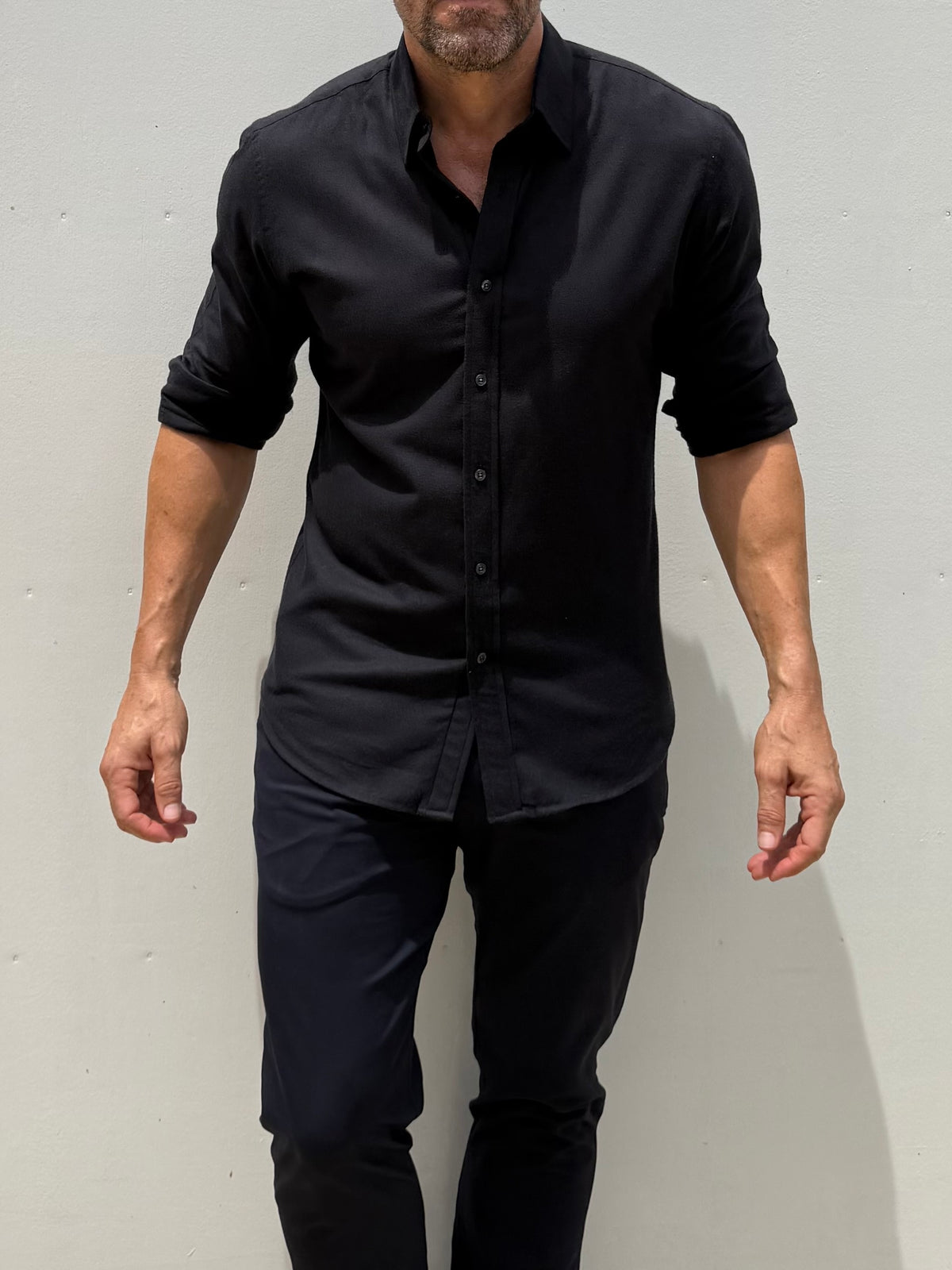 A man wearing a black long-sleeve button-down shirt with rolled-up sleeves and black pants, standing against a light gray wall.