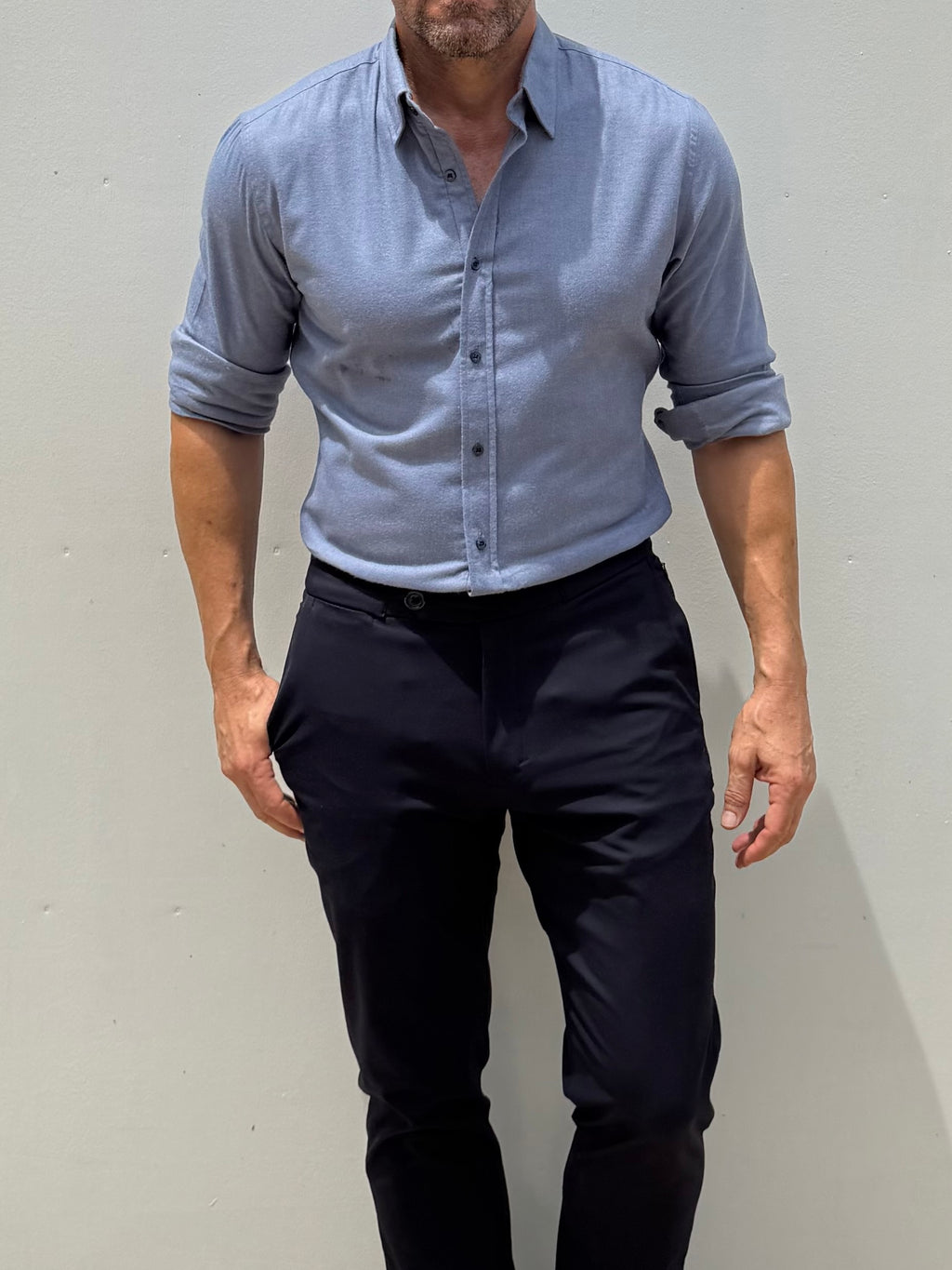 A man wearing a light blue long-sleeve button-down shirt with rolled-up sleeves and dark navy trousers, standing against a plain light gray wall.