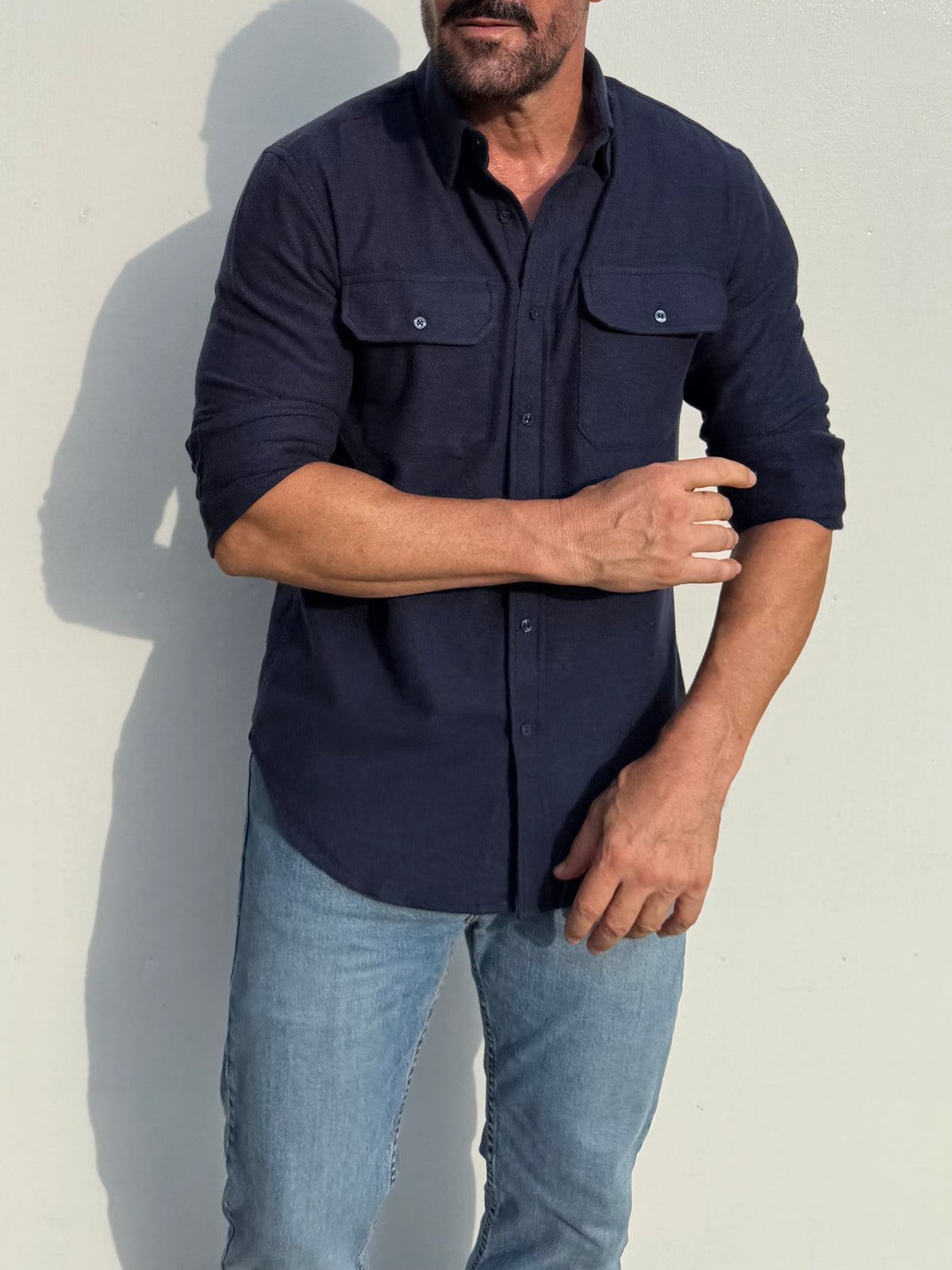 A man wearing a dark navy blue long-sleeve button-down shirt with two chest pockets and light blue jeans, standing against a plain light wall.