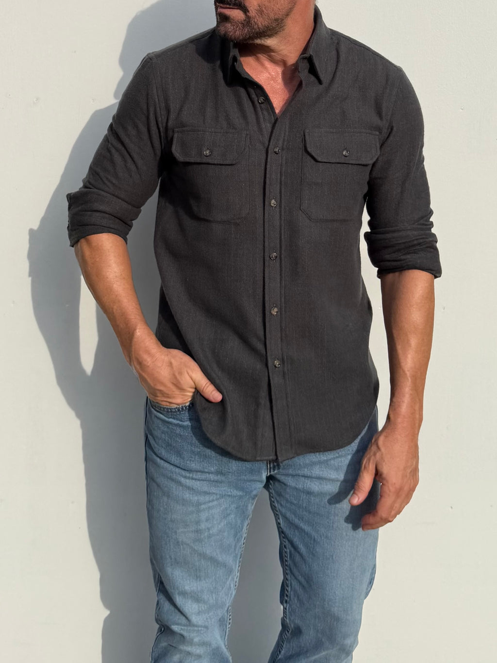 A man wearing a dark charcoal gray long-sleeve button-up shirt with two chest pockets and light blue jeans, standing against a plain light gray wall.