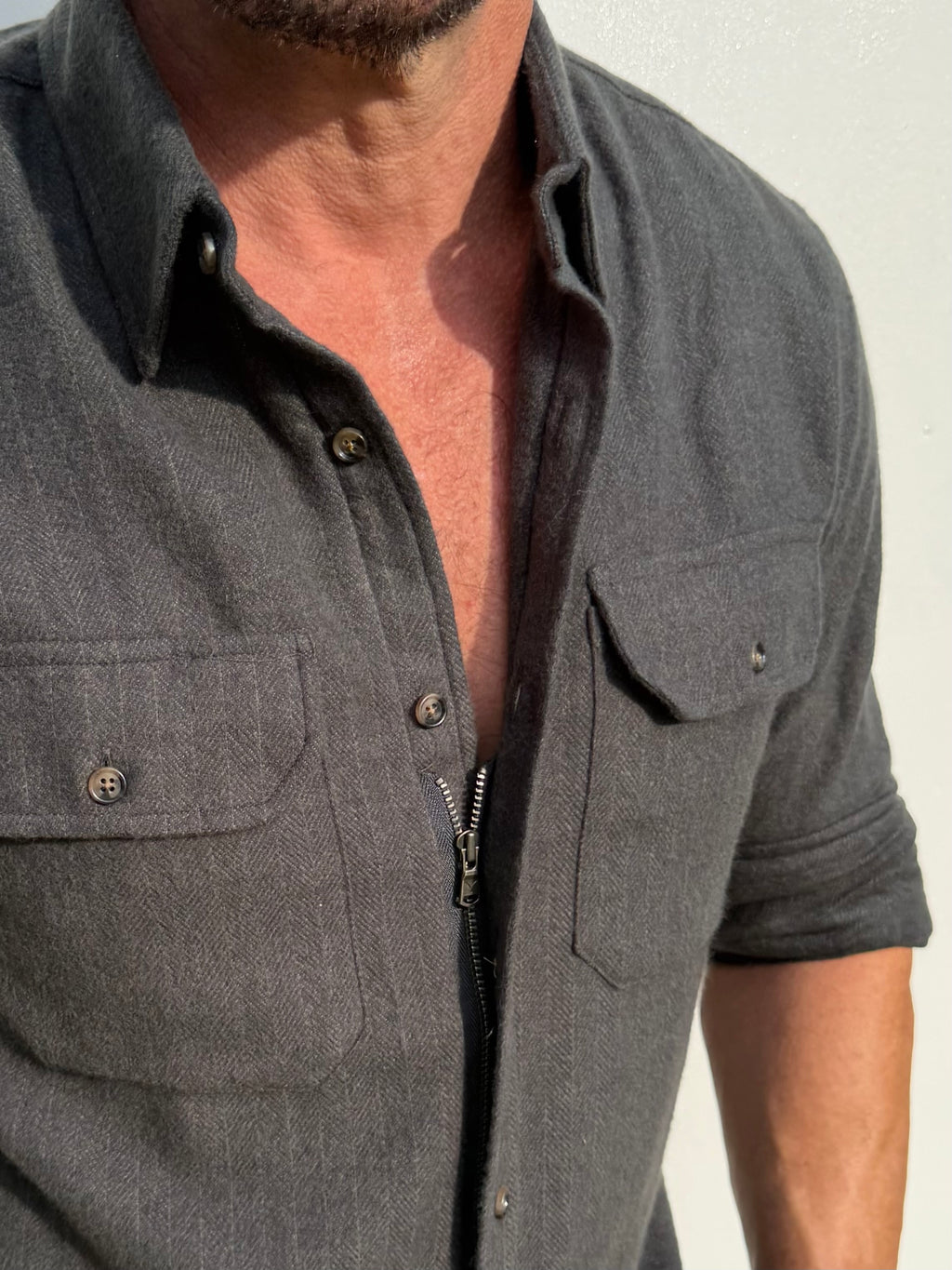 Close-up of a man wearing a dark gray zip-up button-down shirt with two chest pockets and rolled sleeves, set against a plain off-white wall.
