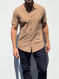 A man wearing a short-sleeve button-up shirt in a warm tan or camel color, paired with dark navy pants, standing against a plain white background.