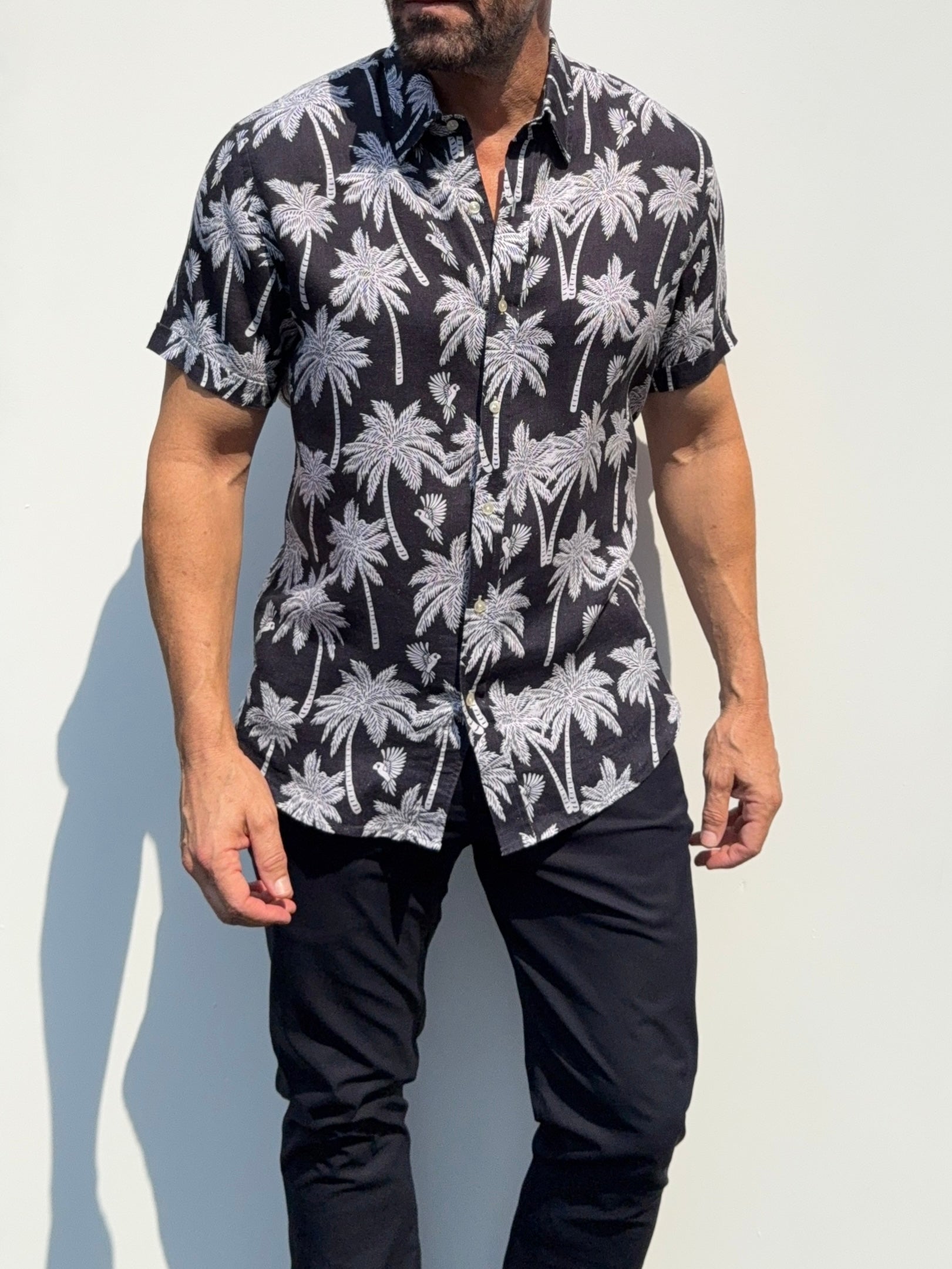 A man wearing a black short-sleeve button-up shirt with a white palm tree pattern, paired with black pants, standing against a light gray wall.