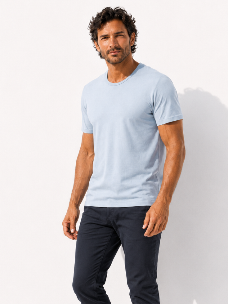 A man modeling a light blue crew-neck t-shirt and dark navy pants, standing barefoot against a white studio background with a soft shadow.