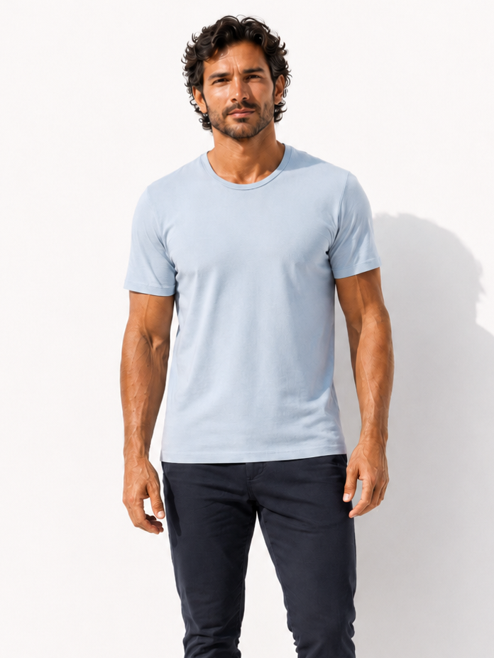 A man modeling a light blue crew-neck t-shirt and dark navy pants, standing barefoot against a clean white studio background with a soft shadow.