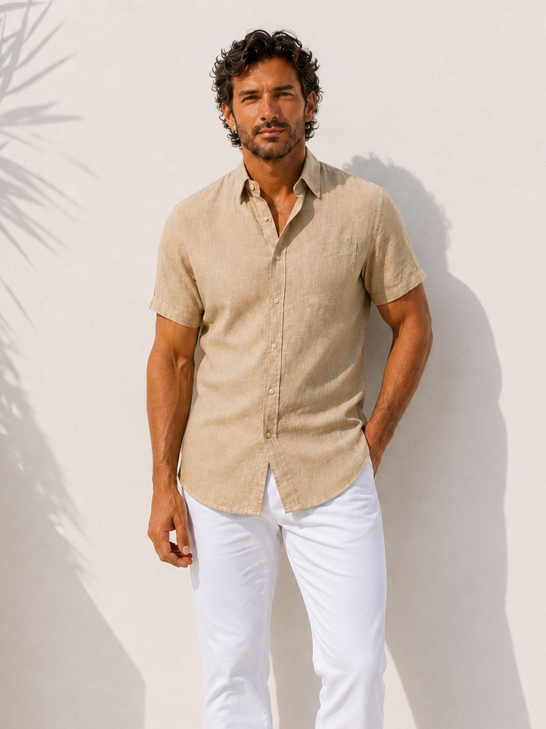 Man wearing a beige shirt and white pants standing against a white wall with a plant to the side.