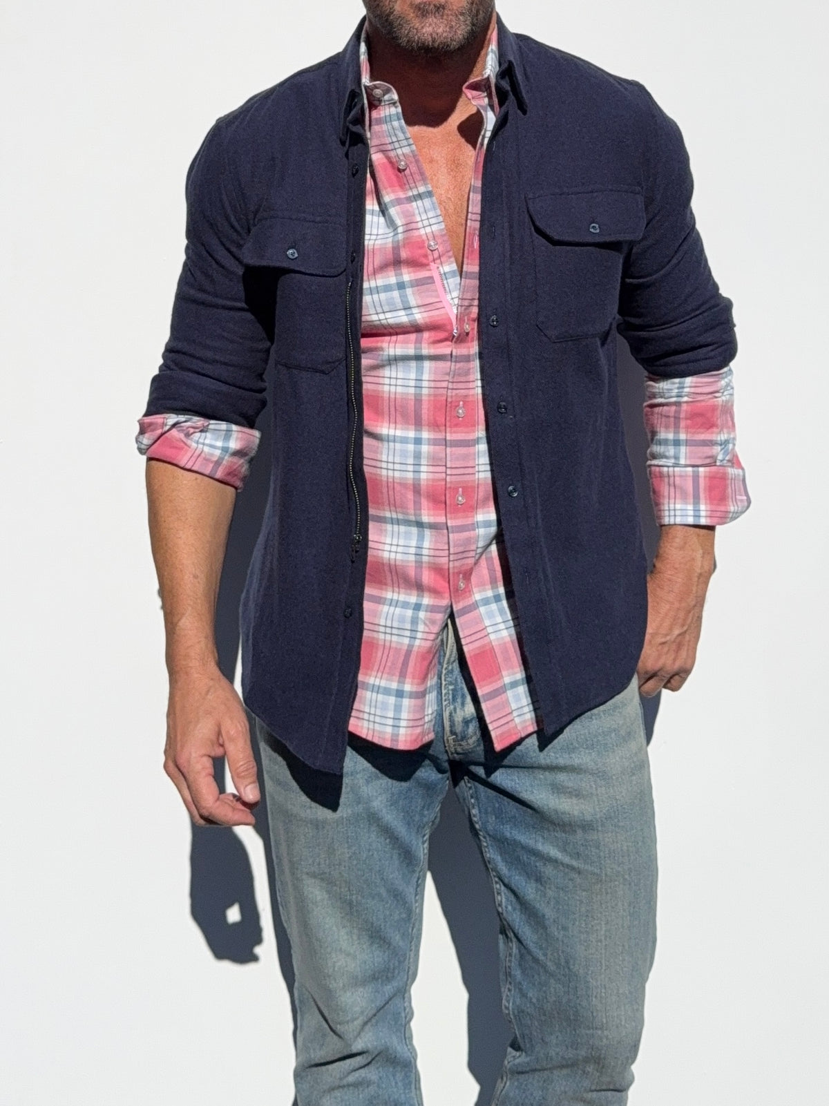 A man wearing a dark navy blue overshirt over a pink and white plaid button-down shirt, paired with blue jeans, shown against a plain white background.