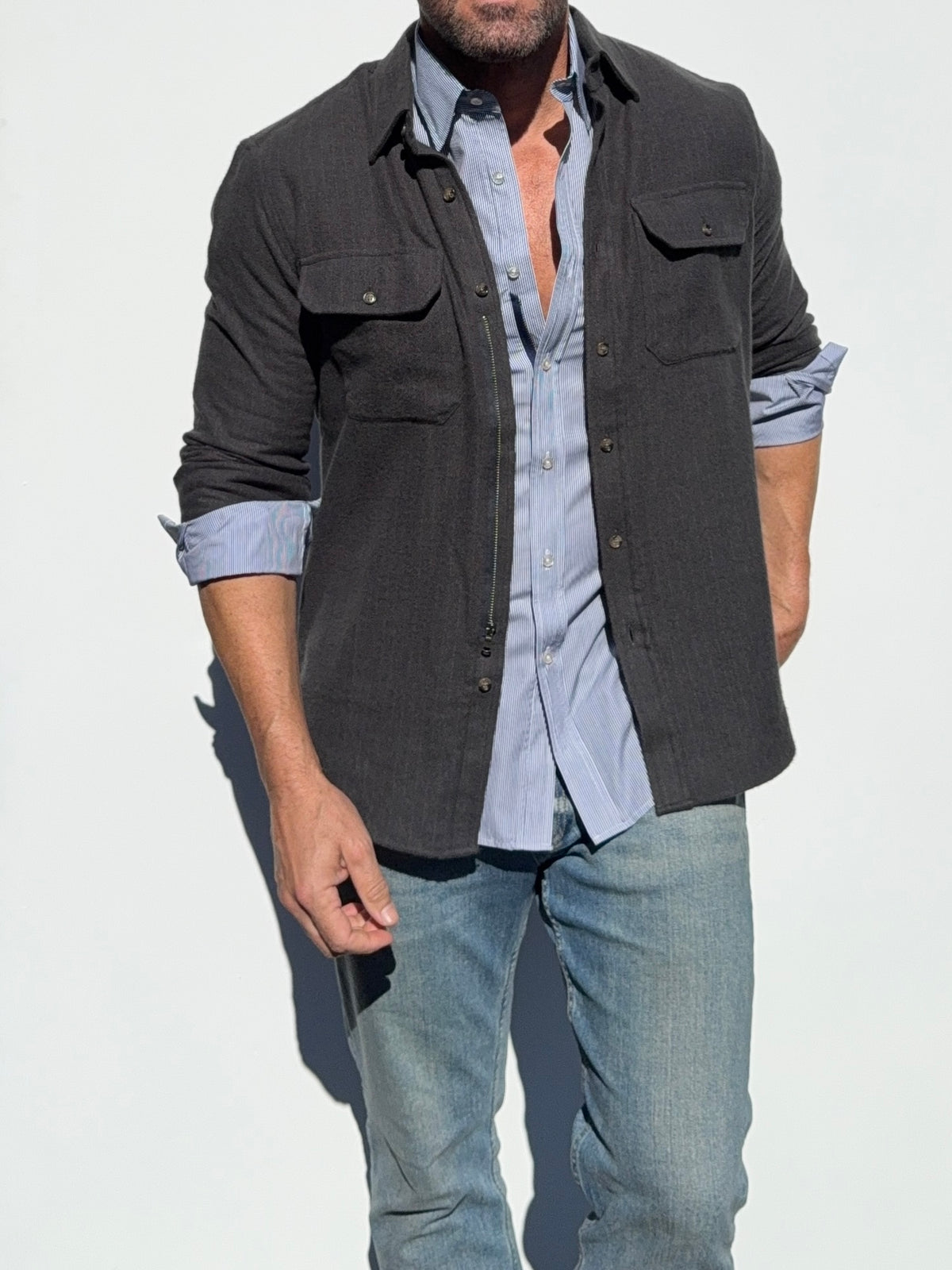 A man wearing a dark charcoal gray button-down overshirt over a light blue button-down shirt and blue jeans, standing against a plain white background.